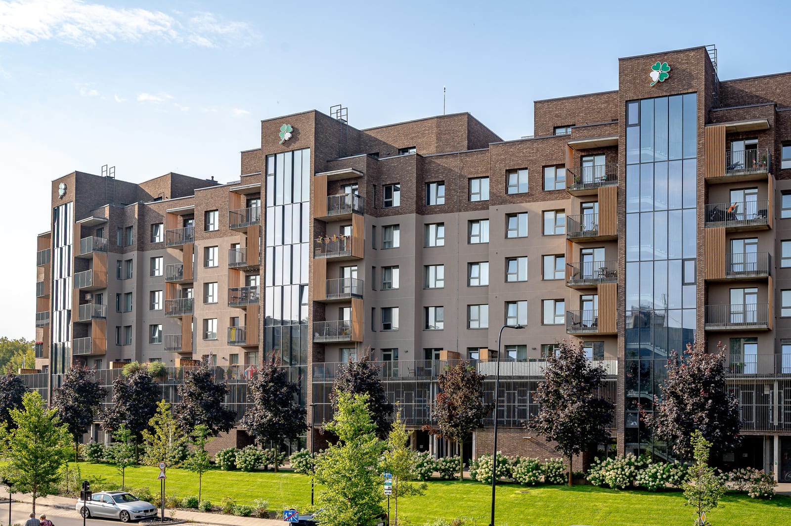 The Lucky Homes complex in Vilnius from the lawn: the full row of brick blocks, young trees and the parking strip